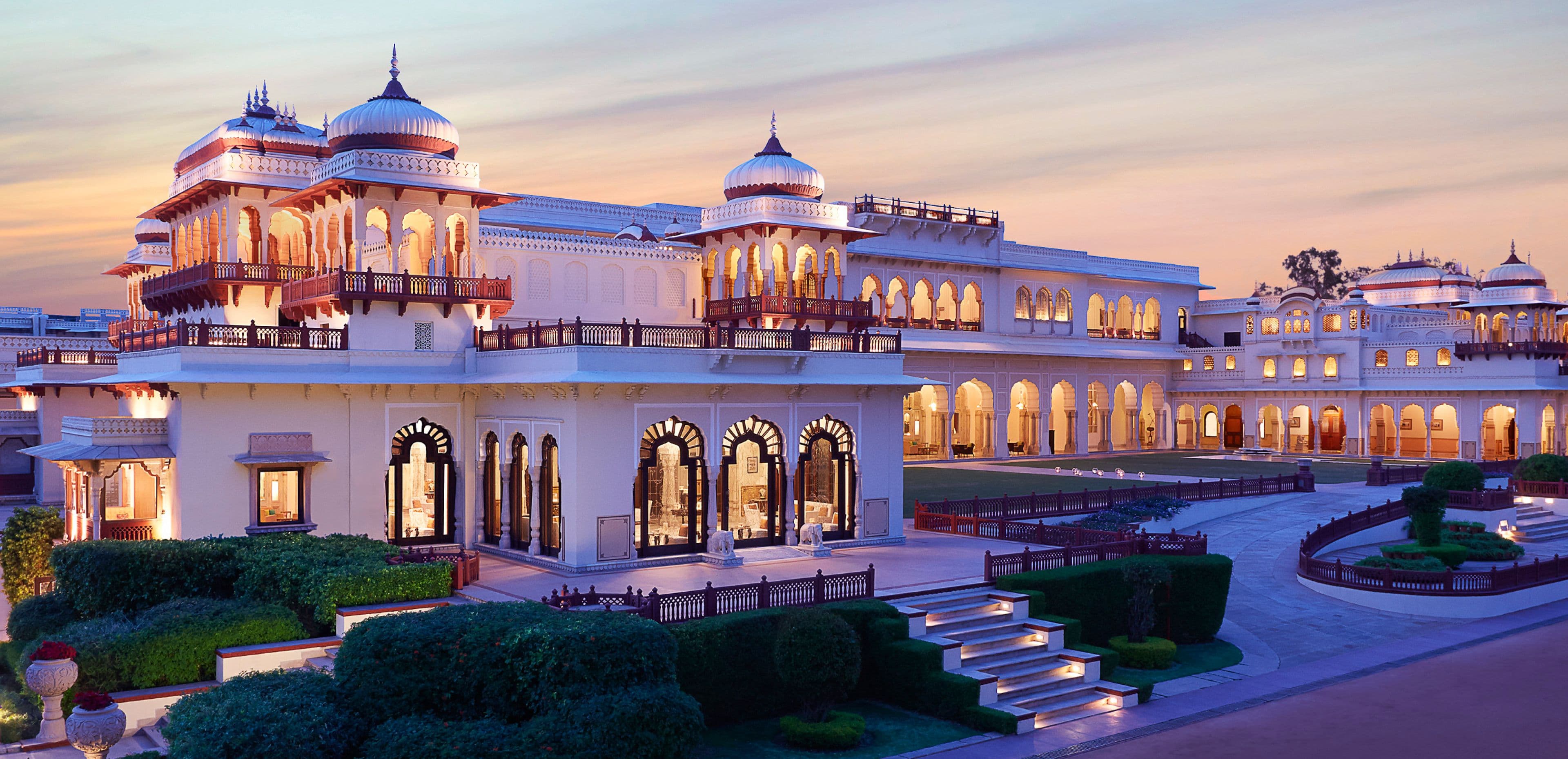 The floodlit sandstone facade and domed cupolas of Rambagh Palace, Jaipur, seen across its formal garden