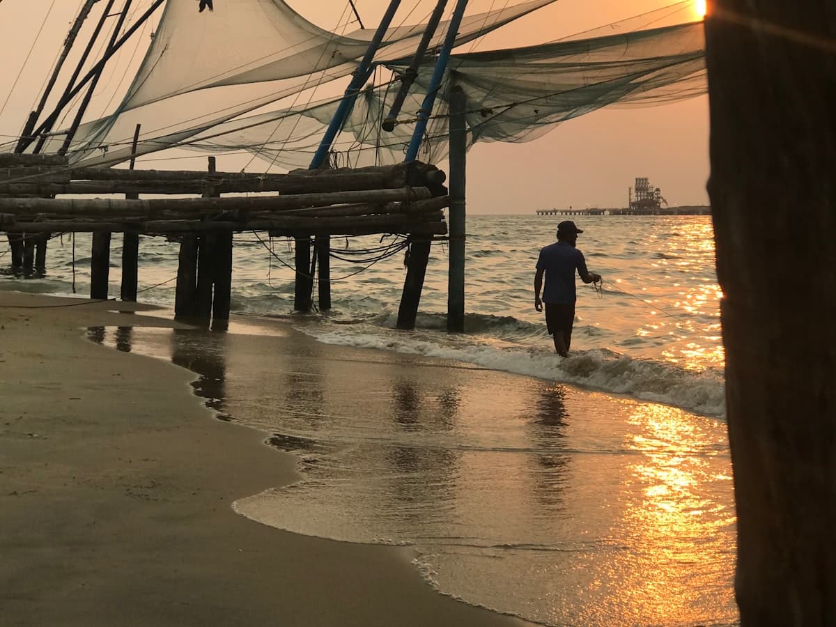 A man walking along the shoreline beside a cantilevered Chinese fishing net at Fort Kochi