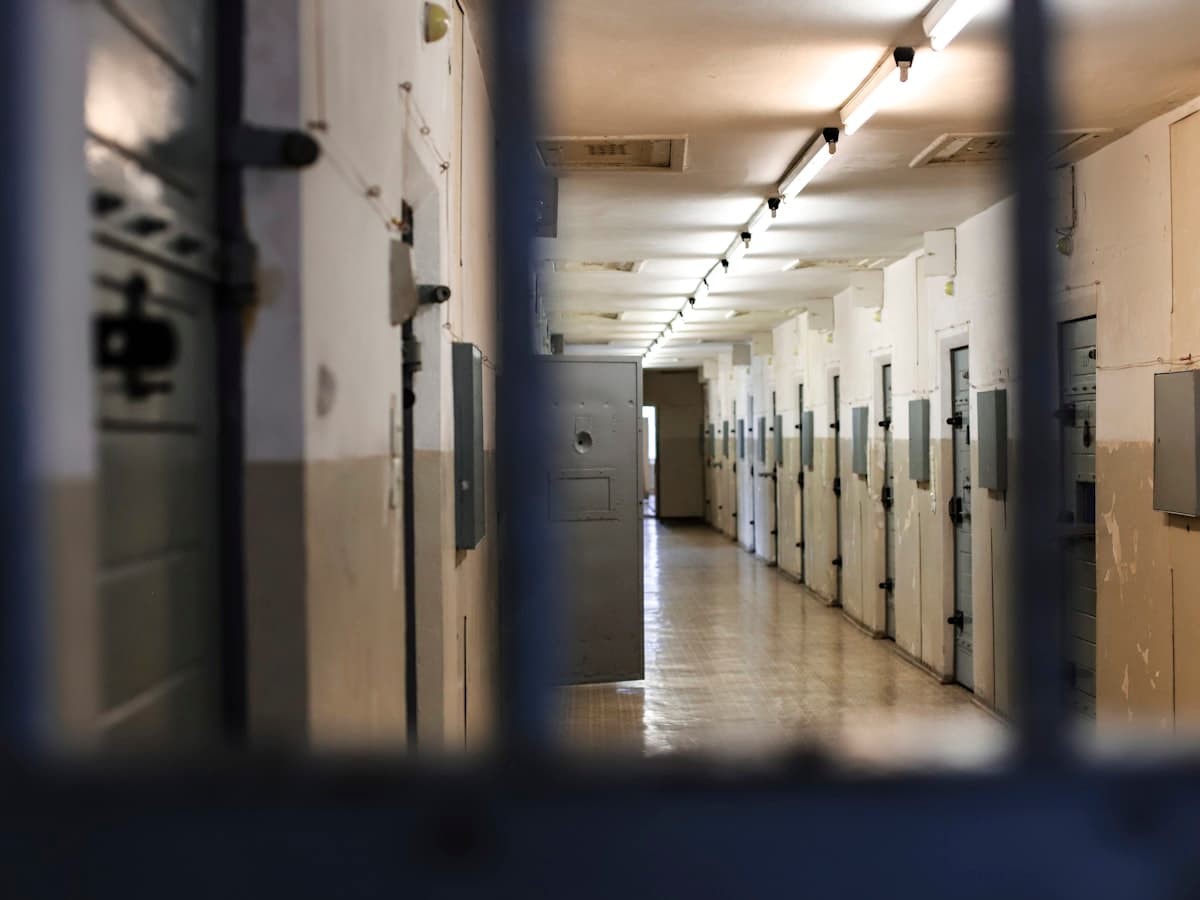A long prison corridor with rows of metal doors seen through dark bars